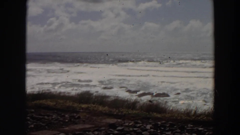 1964 California 8mm coastline frame with churning waves, low grass, rocks, and a cloudy horizon.