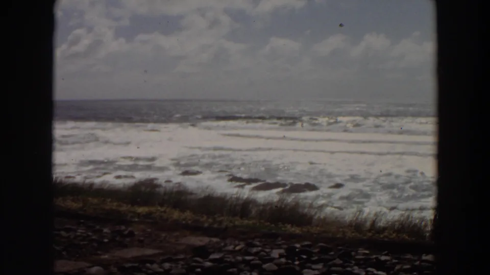 1964 California 8mm view of foamy ocean water reaching a rugged shore with grass and stones.