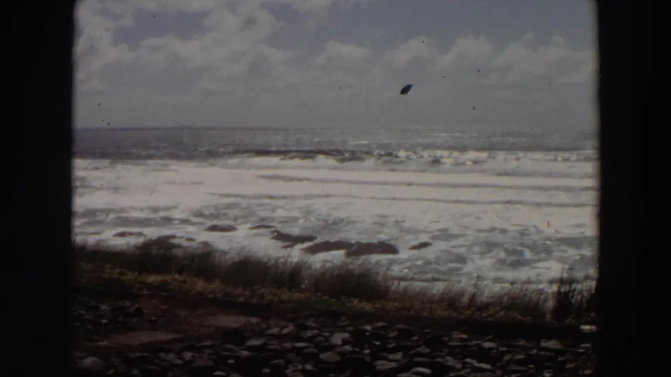 1964 California coastal surf rolls toward a rocky shoreline with grass and a partly cloudy sky, seen through a dark foreground frame.