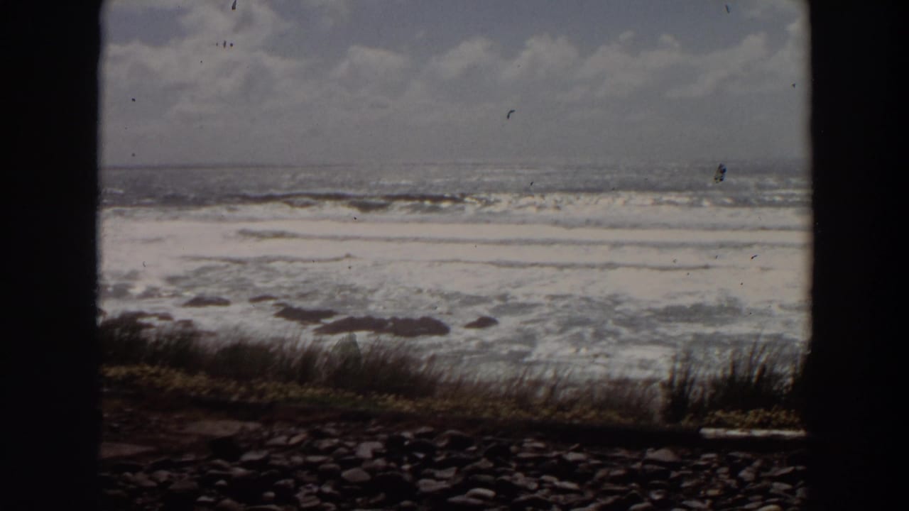 A bright California ocean view shows white surf, scattered rocks, and cliff-edge grass within dark 8mm frame borders.