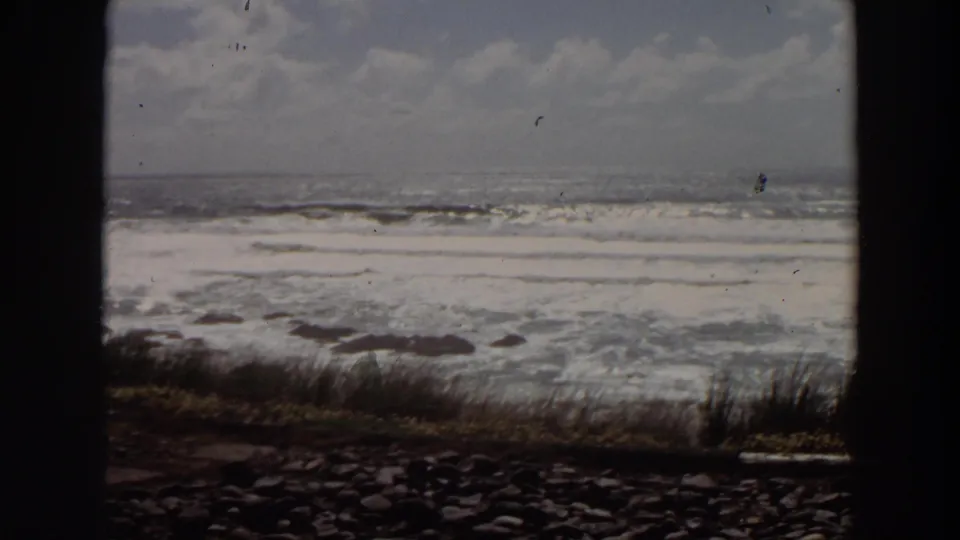 A bright California ocean view shows white surf, scattered rocks, and cliff-edge grass within dark 8mm frame borders.