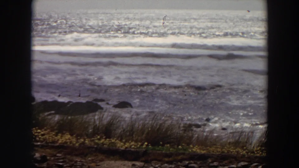 1964 California shoreline frame with churning surf, dark rocks, coastal grass, and visible 8mm grain and scratches.