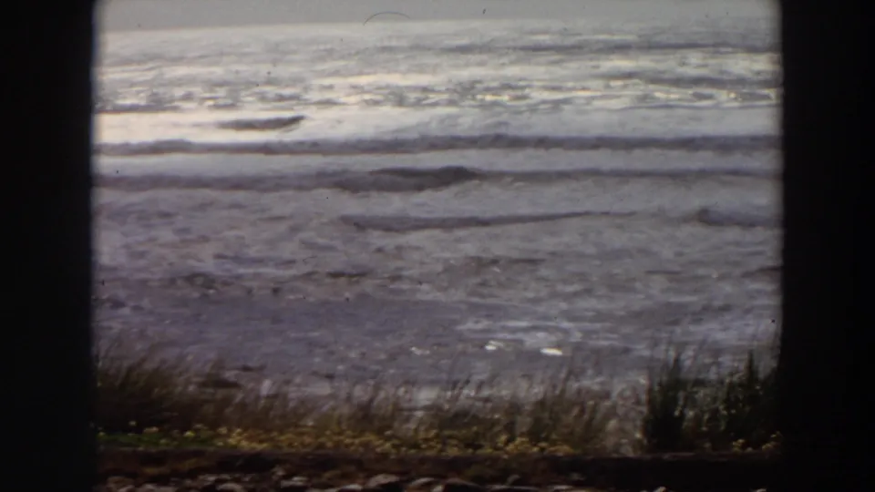 Vintage 8mm frame of a California beach, showing pale surf lines, wet sand, foreground grasses, and dark border areas from the film frame.