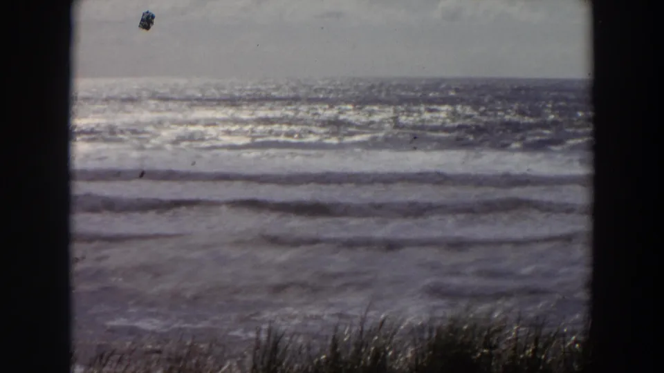 1964 California 8mm view of ocean waves rolling toward shore, seen past beach grass and dark vertical framing.