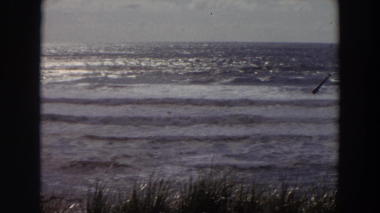1964 California coastal view with bright surf, low horizon, beach grass, and dark frame edges from the 8mm scan.
