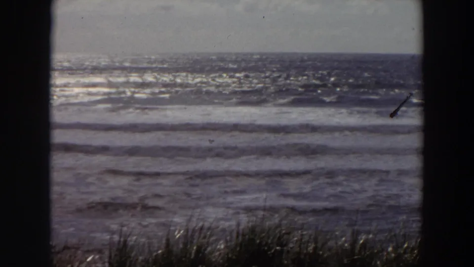 1964 California coastal view with bright surf, low horizon, beach grass, and dark frame edges from the 8mm scan.