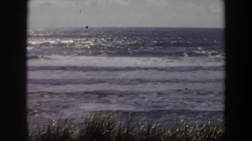 Vintage 8mm California shoreline frame showing choppy ocean, rolling surf, and beach grass in the foreground.
