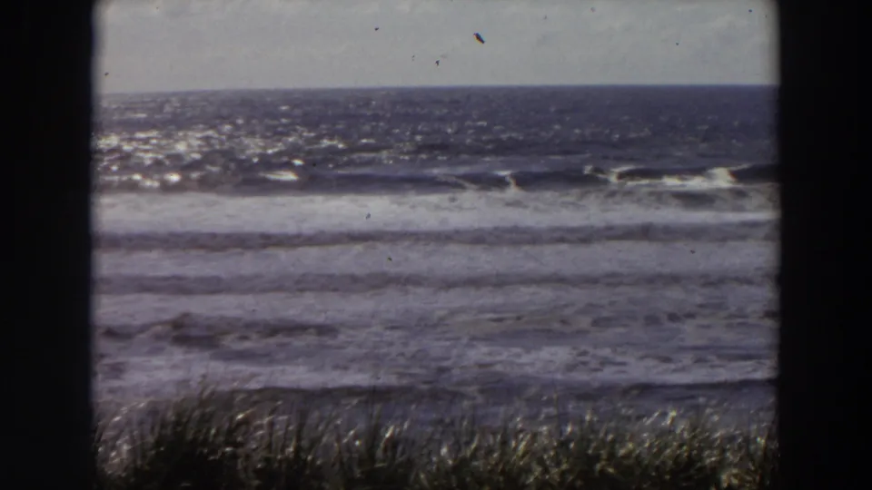 1964 California home-movie frame of ocean waves and beach grass, with dark vertical edges framing the view.
