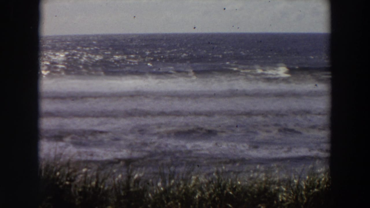 1964 California coastal home-movie frame showing surf, horizon, grasses, and dark window-frame borders, with no person clearly centered.