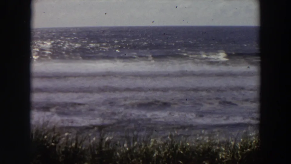 1964 California coastal home-movie frame showing surf, horizon, grasses, and dark window-frame borders, with no person clearly centered.