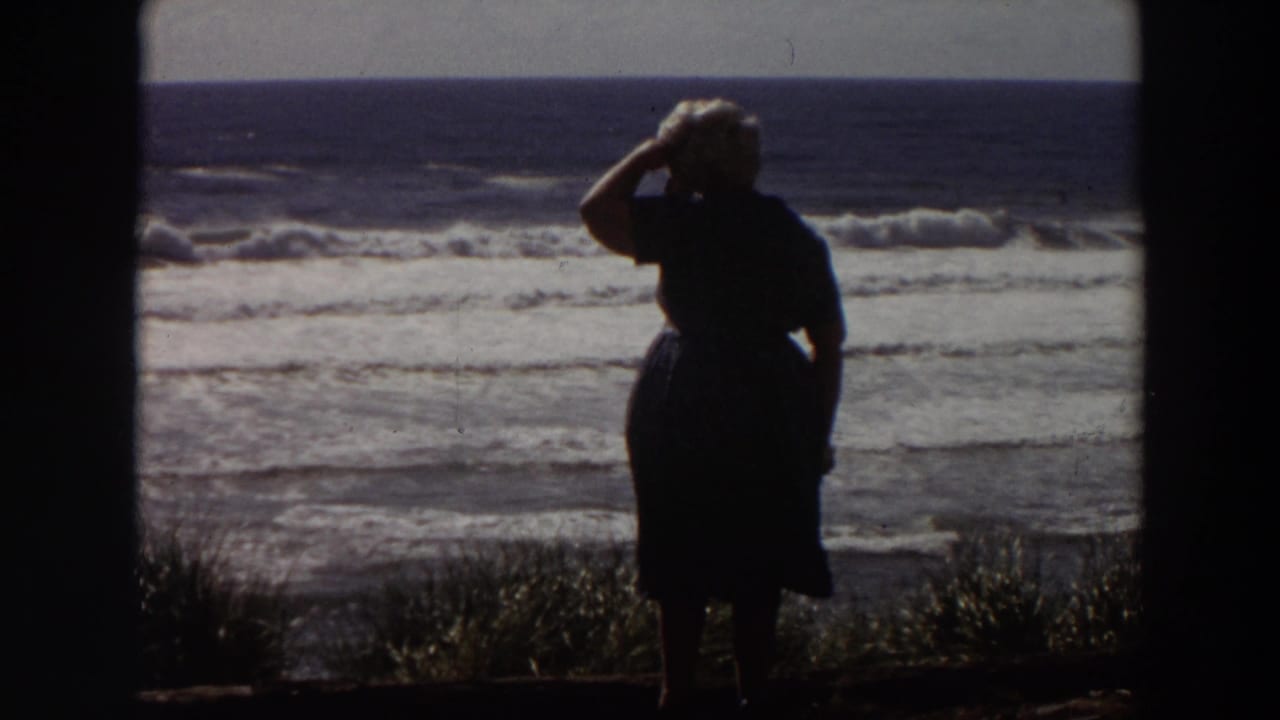 Silhouetted woman stands near the coast, raising a hand to shield her eyes from the sun.
