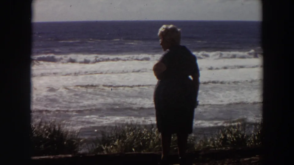 1964 California home-movie frame showing a silhouetted woman at a coastal overlook, with white surf and ocean horizon in the background.