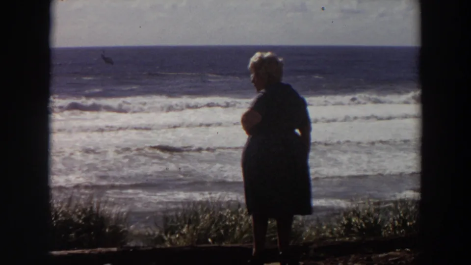 1964 California coastal frame with a silhouetted woman watching the surf, framed by grass in the foreground and open ocean beyond.