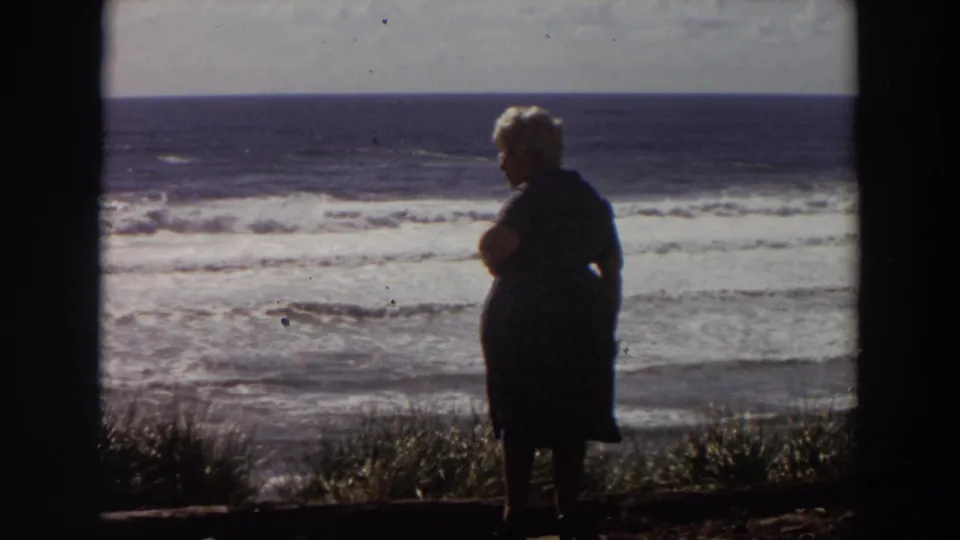 Elderly woman in a dark dress stands at a California coastal overlook in 1964 home-movie footage, facing the surf.