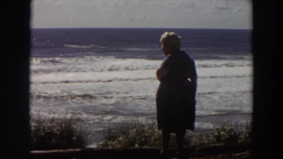 A silhouetted older woman stands on a grassy California bluff, with layered surf visible in soft 8mm color.