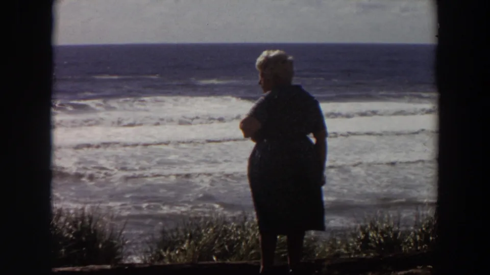 Near the end of the clip, the older woman remains by the coastal grass as waves roll in behind her in 1964 California footage.