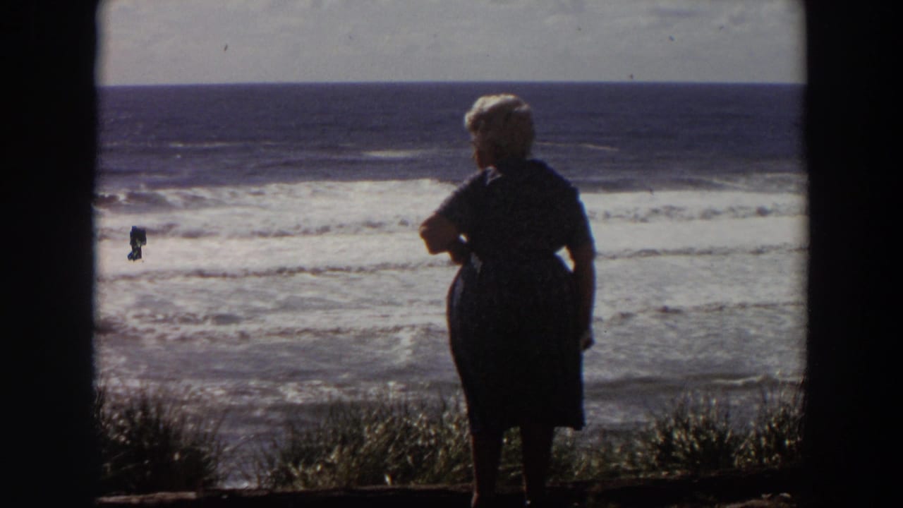 Older woman in dark clothing stands near the California coastline in 1964, silhouetted against surf and bright sky.