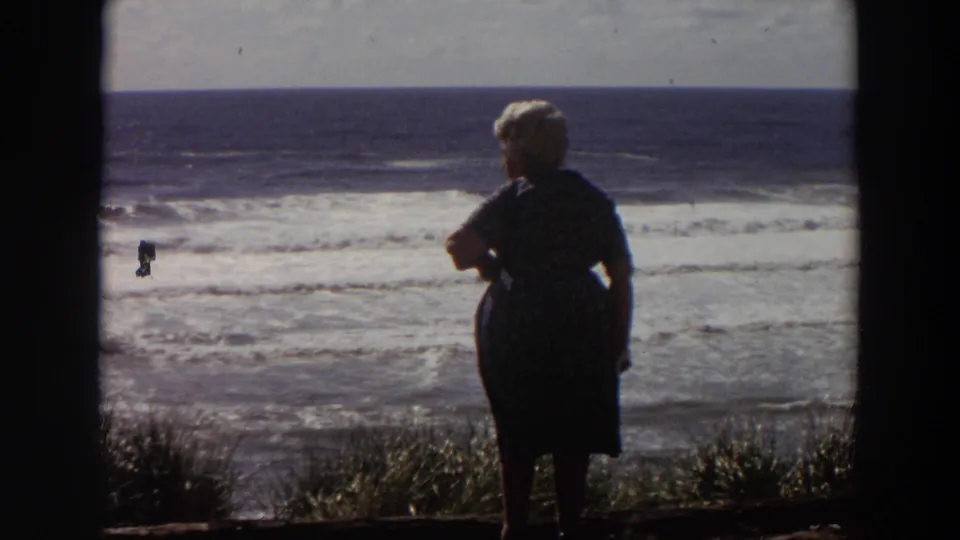 Older woman in dark clothing stands near the California coastline in 1964, silhouetted against surf and bright sky.