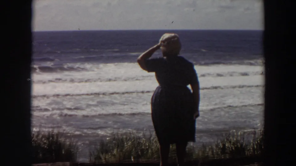 Older woman shades her eyes while looking toward the Pacific surf, framed by dark 8mm gate edges.