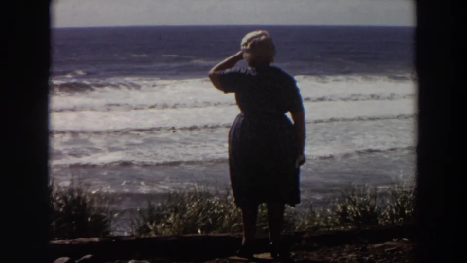 A silhouetted older woman stands at a California shore viewpoint in 1964, with layered surf visible behind her.
