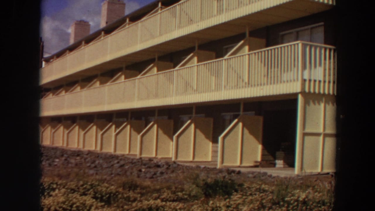 1964 California home-movie frame showing a pale apartment building with long balconies, chimneys, and rocky foreground planting.