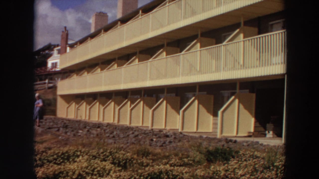 Apartment exterior with long balcony railings and a woman partly visible near the slope.