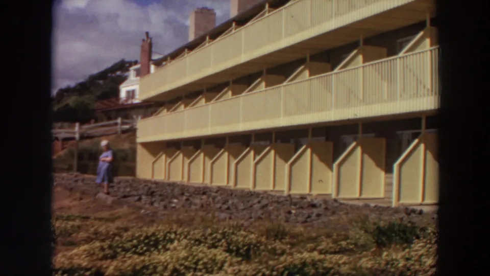 1964 California home-movie frame showing a woman in a light dress standing on rocky ground below a multi-story apartment building.