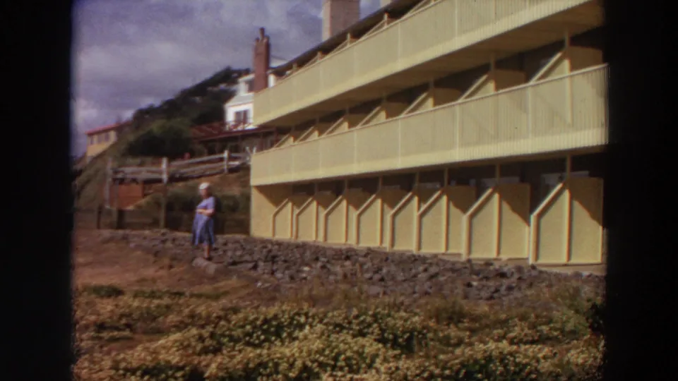 1964 California frame of a woman standing on a rocky landscaped slope beside a pale multi-story apartment exterior with balconies.