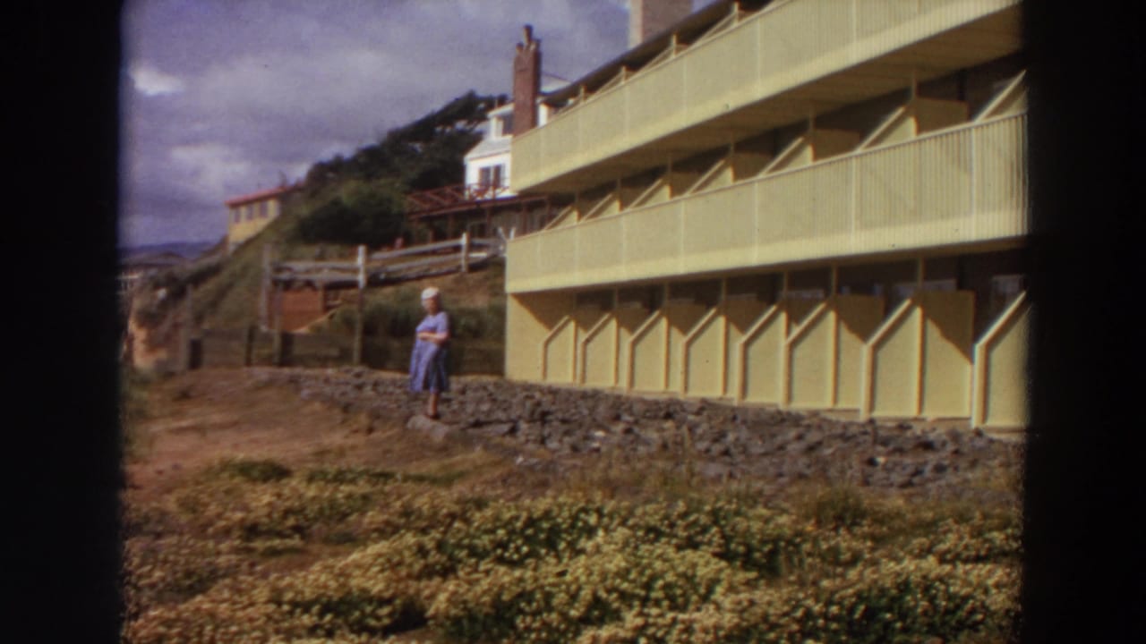 Woman in a blue dress stands near a yellow coastal apartment building above a grassy slope.