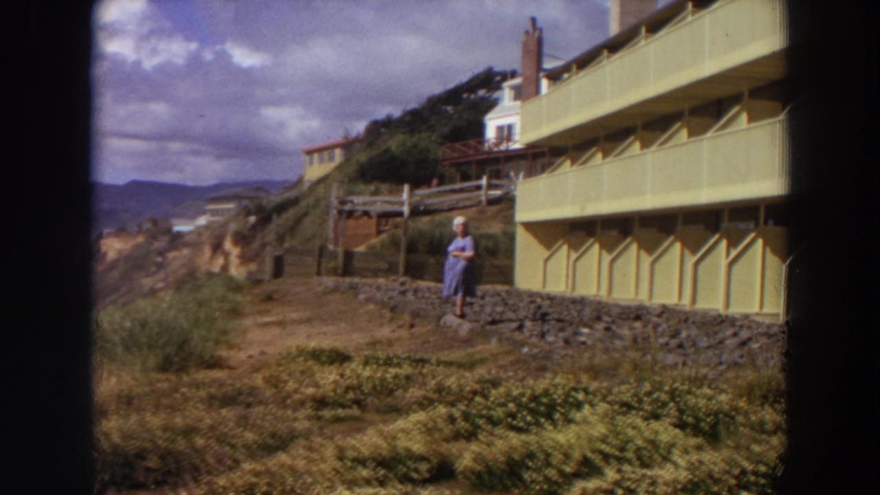 Woman stands outside the yellow apartment as the view opens toward cliffs and distant hills.