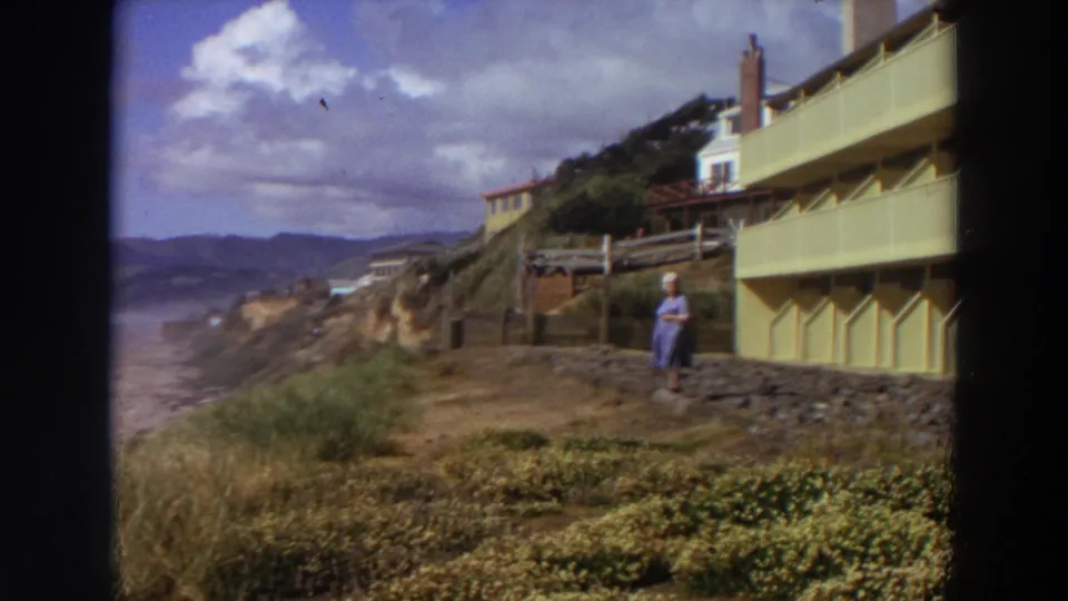 A 1964 California coastal frame shows ocean surf and cliffs at left, with the woman still near the yellow apartment at right.