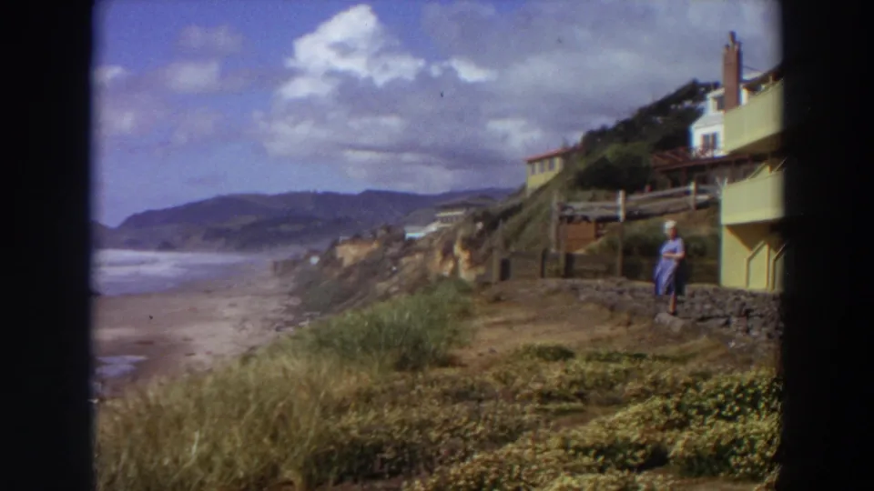 The final frame widens to the California coastline, keeping the apartment edge and woman near the right side of the 8mm home-movie view.