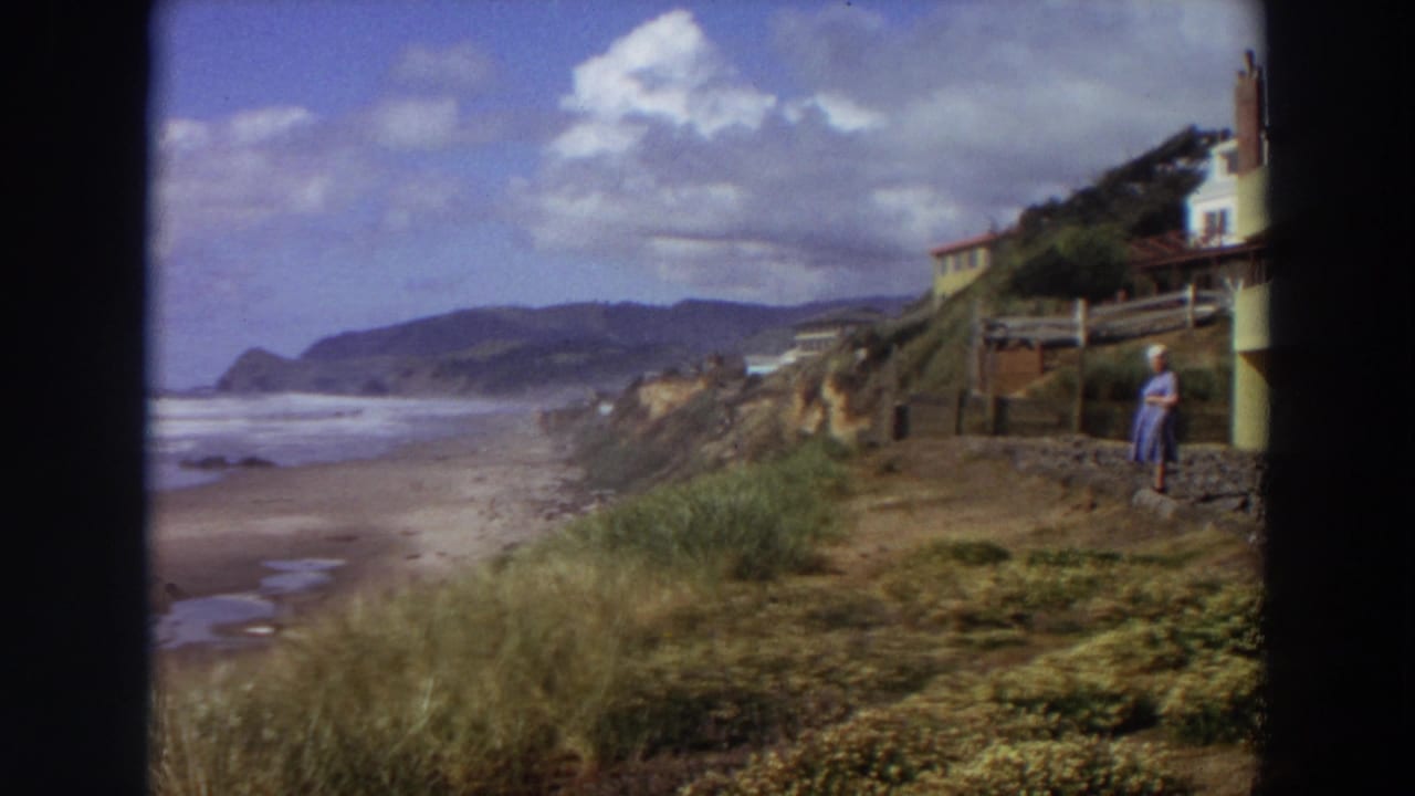 Coastal beach view with surf, grassy foreground, cliff houses, and a woman near a path.