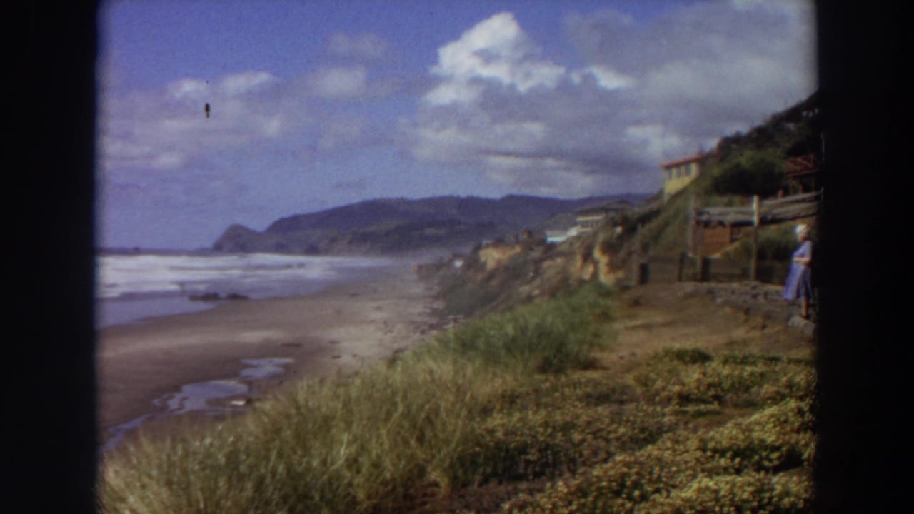1964 California shoreline view with waves, beach, grassy bluff, cliff houses, and a partly framed woman at right.