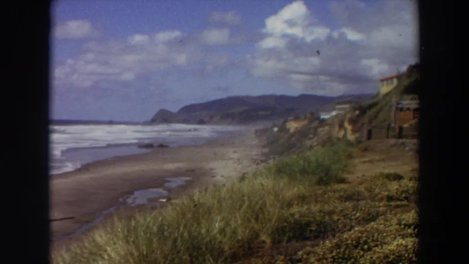 1964 California coast frame with beach surf, foreground grasses, cliffside structures, and a hazy headland in the distance.