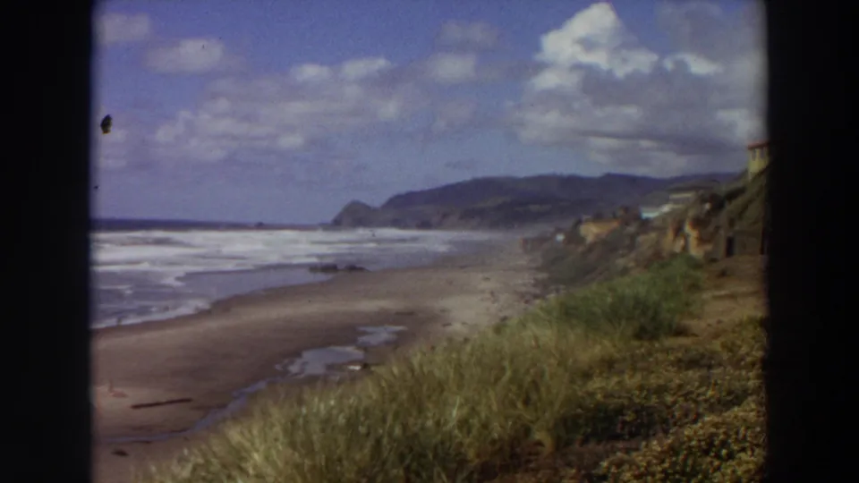 1964 California coastal home-movie frame showing waves, sand, grass-covered bluff, cliff houses, and distant hills.