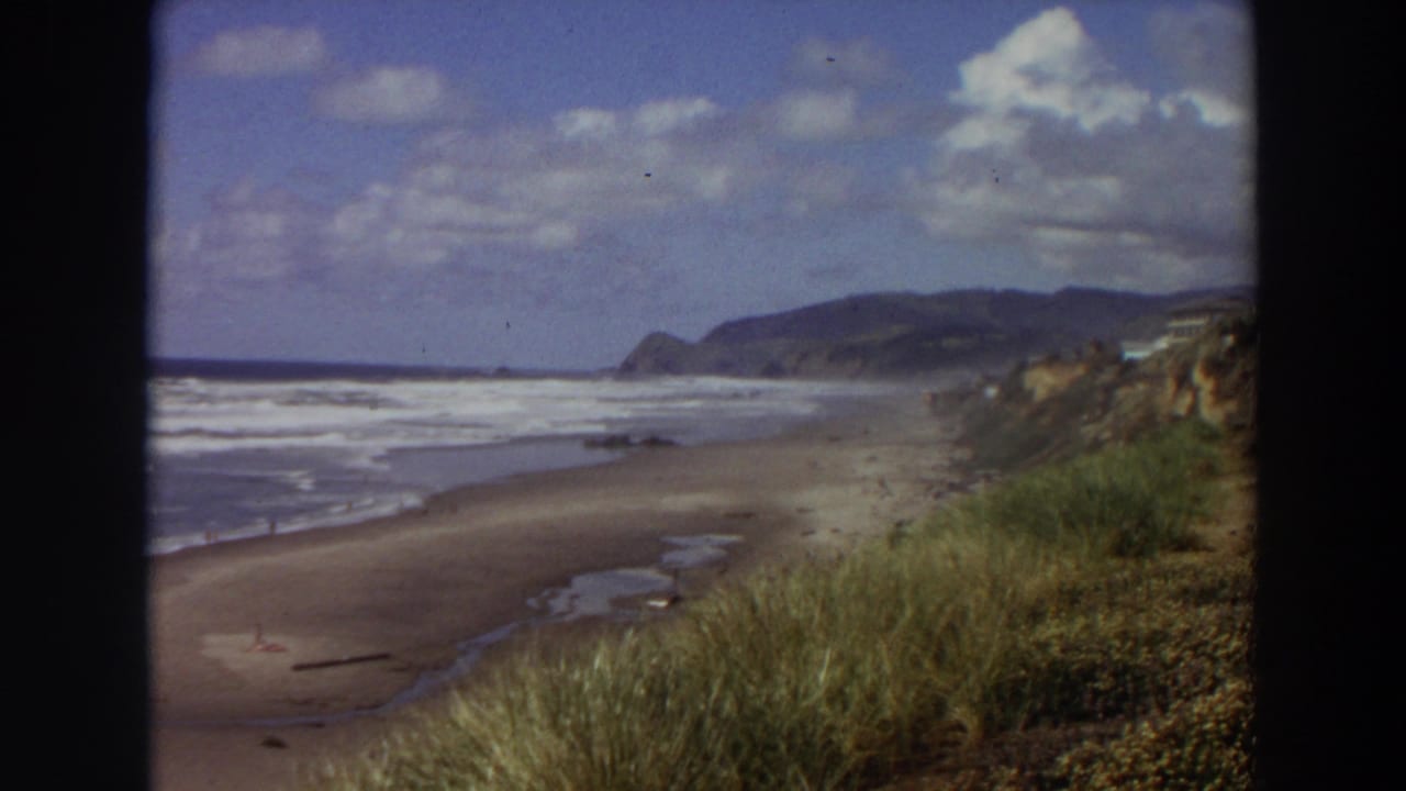 1964 California 8mm view of surf rolling onto a broad sandy beach, with dune grass in the foreground and coastal hills in the distance.
