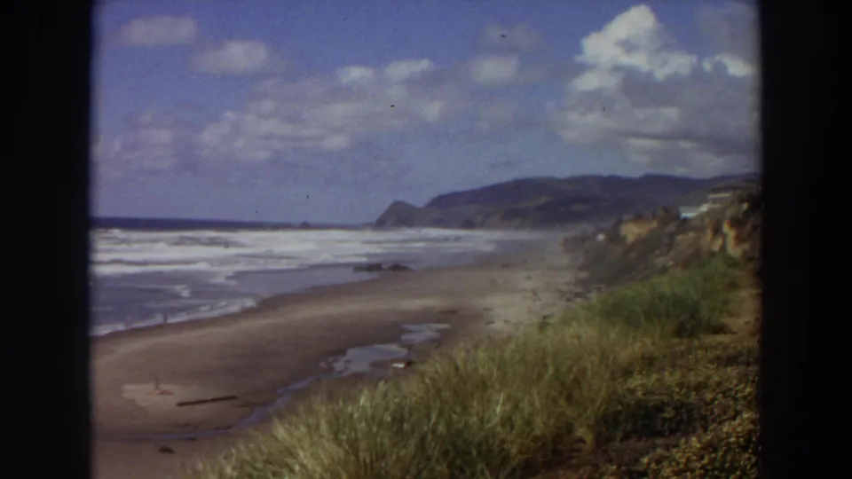 1964 California 8mm view of surf rolling onto a broad sandy beach, with dune grass in the foreground and coastal hills in the distance.