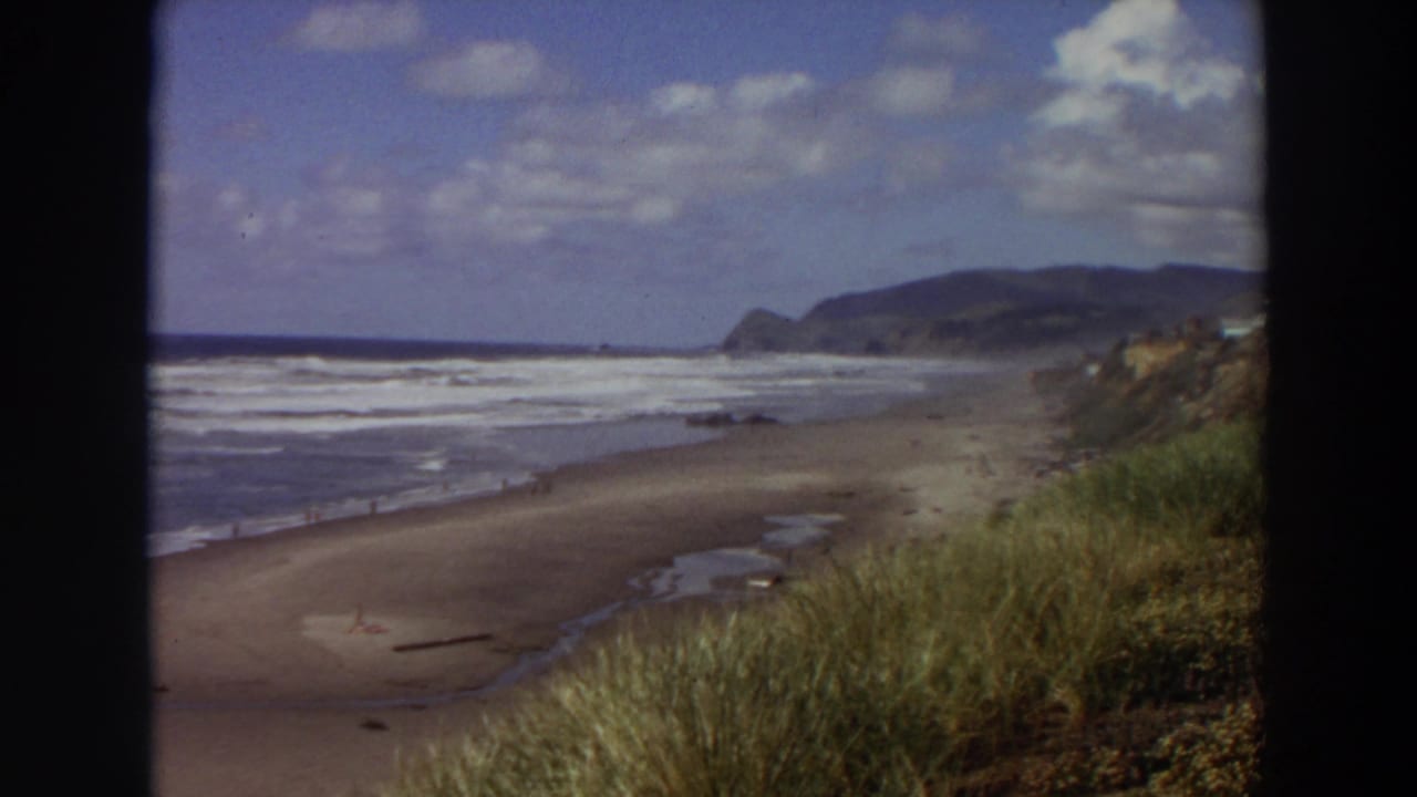 Surf line, wide beach, grassy dunes, distant headland, and blue sky with clouds.