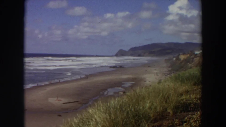 1964 California coastal home-movie frame showing a broad surf line, sandy beach, grassy dune edge, and distant hills under scattered clouds.