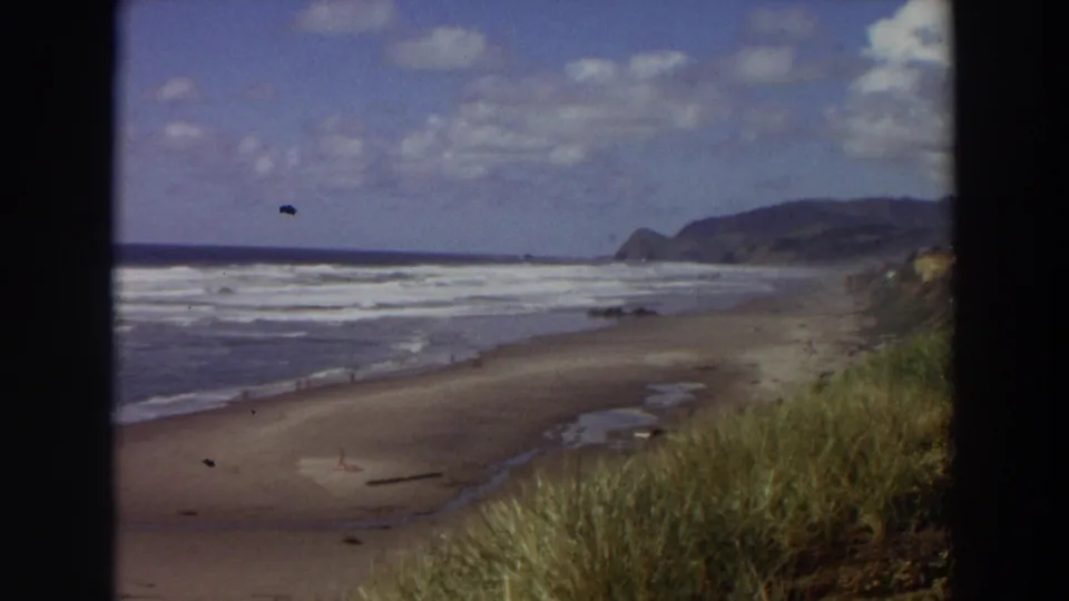 1964 California 8mm frame of waves breaking along the shoreline, with tall dune grass close to camera and tiny beachgoers near the water.