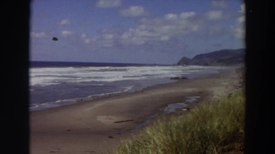 1964 California home-movie frame with surf crossing a wide beach, dune grass in the foreground, distant coastal hills, and visible film specks.