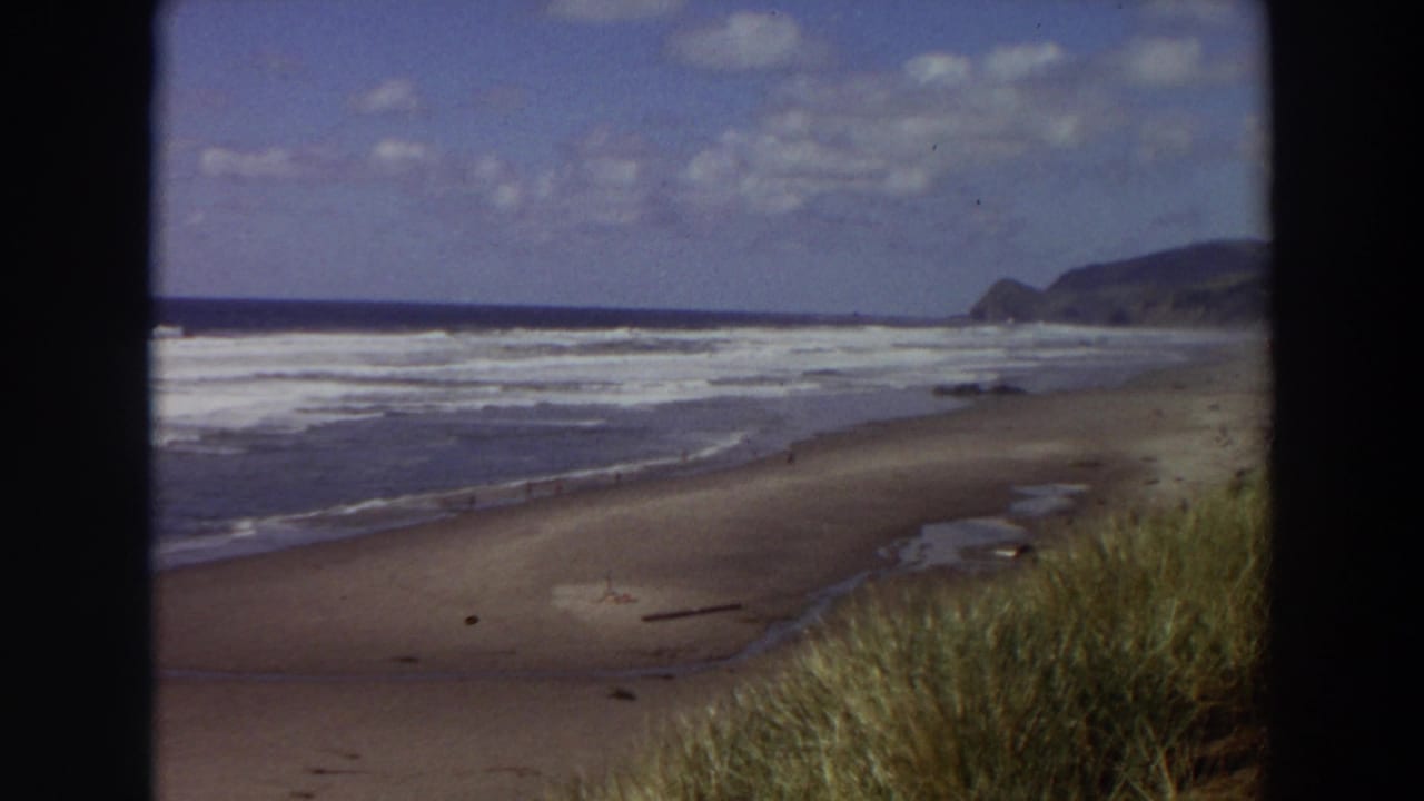 Wide sandy beach with rolling waves, beach grass, distant headland, and partly cloudy sky.