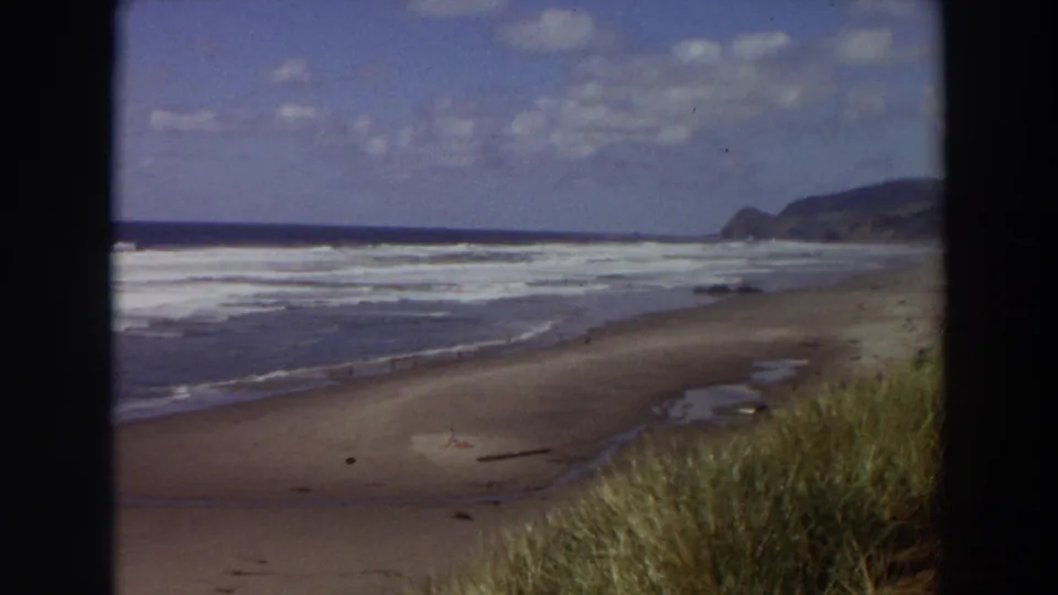 1964 California 8mm frame of a broad beach, white surf, dune grass in the foreground, and distant coastal hills under broken clouds.
