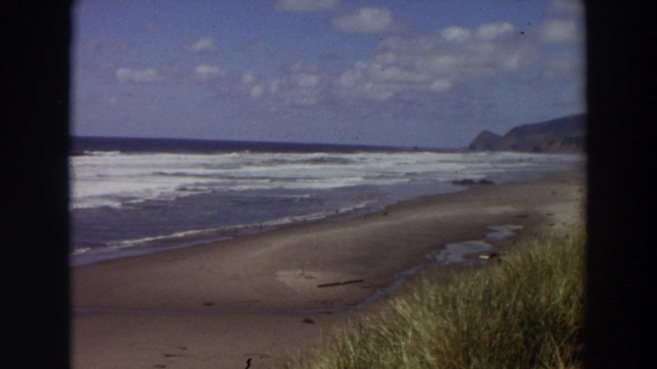 1964 California shoreline view on 8mm film, showing low beach grass, wet sand, repeated surf bands, and a dark headland in the distance.