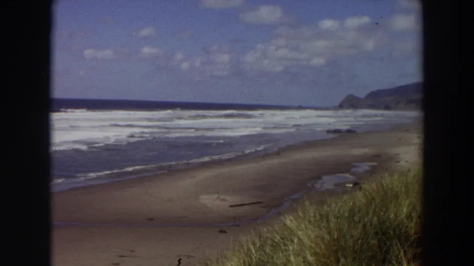 1964 California shoreline view on 8mm film, showing low beach grass, wet sand, repeated surf bands, and a dark headland in the distance.