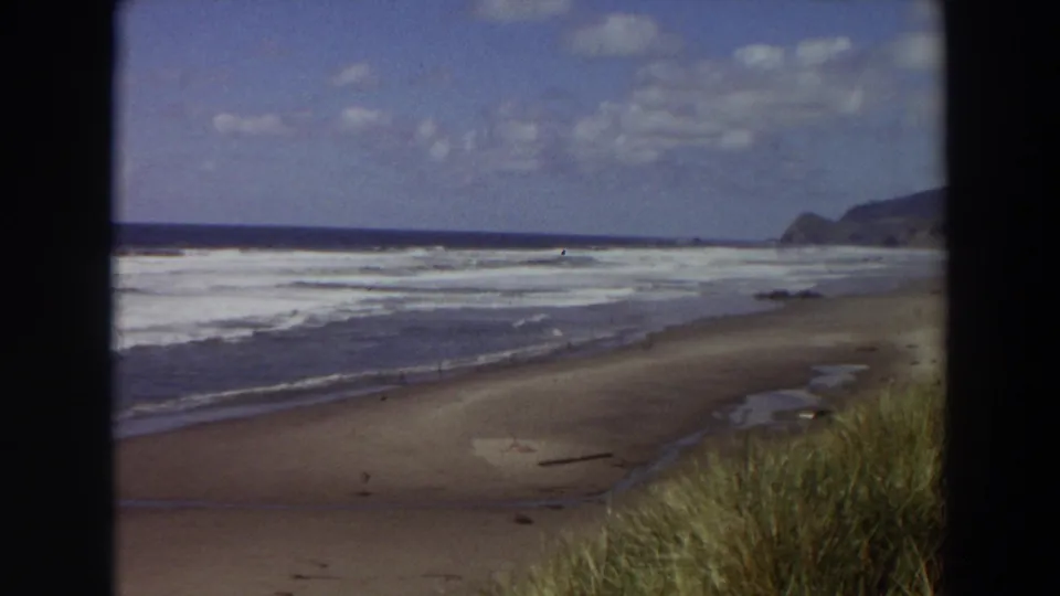 1964 California 8mm coastal frame with a quiet sand beach, foamy waves, wind-shaped grass, and a distant headland softened by film grain.