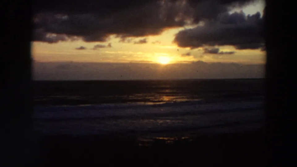 1964 California 8mm sunset or sunrise frame with the sun near the horizon, dark cloud cover, ocean reflections, and a distant beach figure.