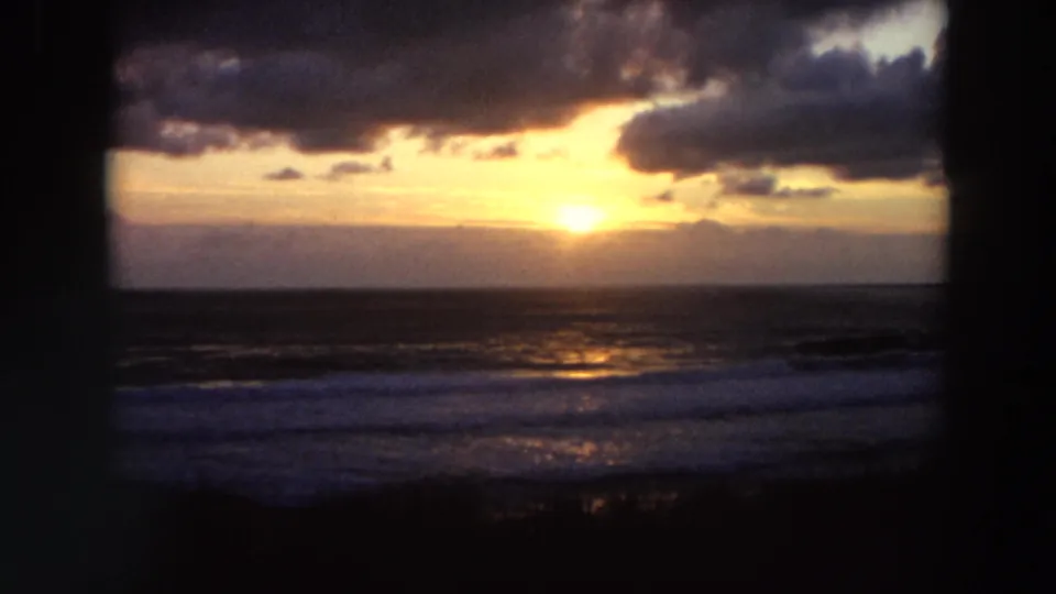 1964 California coastal sunset seen through dark framing, with the sun glowing above the horizon and light reflecting across rolling surf.