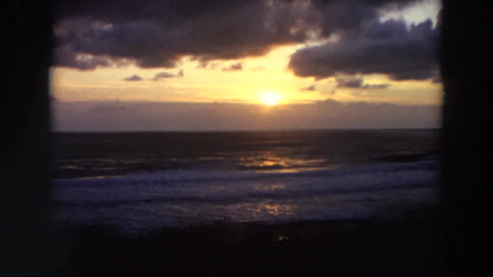 1964 California 8mm frame showing a bright sunset partly veiled by clouds, ocean horizon, shimmering water, and surf below.